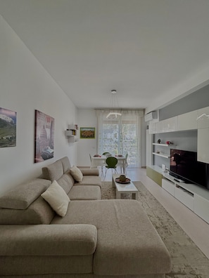 Ein modernes Wohnzimmer mit einer Couch, einem Esstisch mit Stühlen, einem Fernseher und einem großen Fenster.