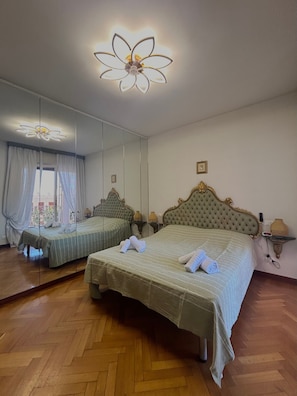 Una habitación de hotel con dos camas, un espejo grande, un ventilador de techo y suelo de madera en patrón espiga.