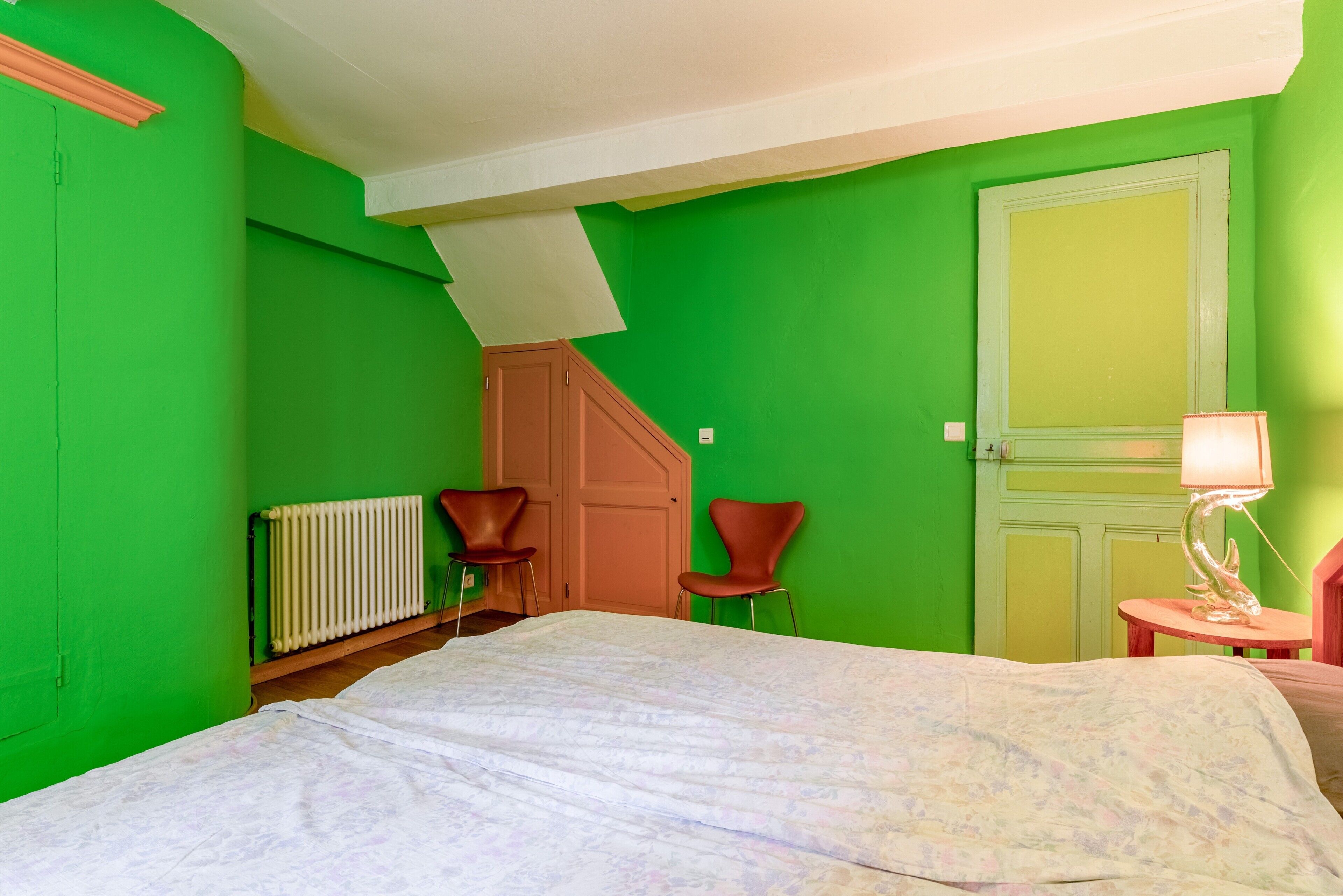 Quarto com uma cama, duas cadeiras, um radiador, uma porta e um abajur.