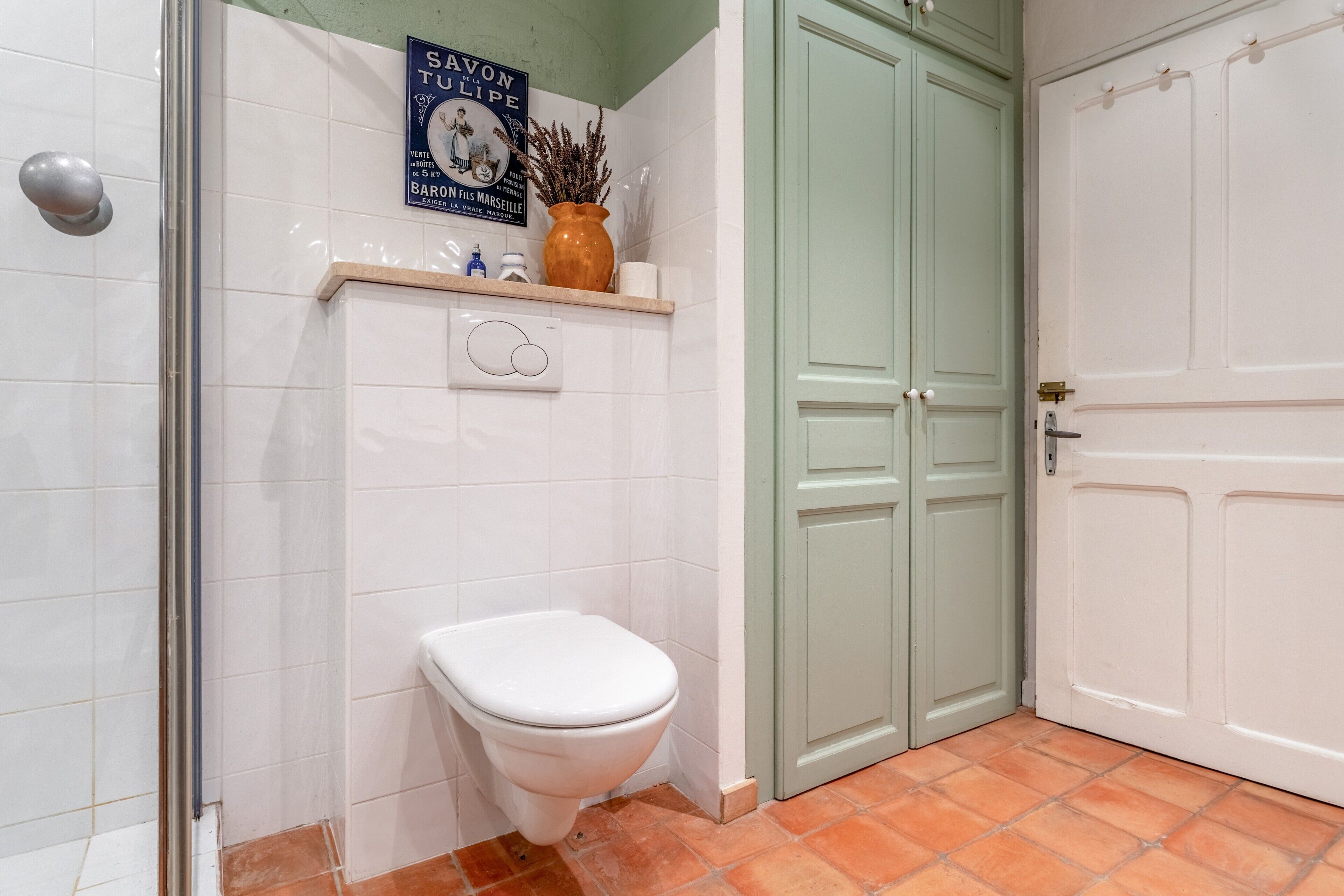 Un baño con inodoro blanco, ducha, puerta verde y estante con artículos de aseo.