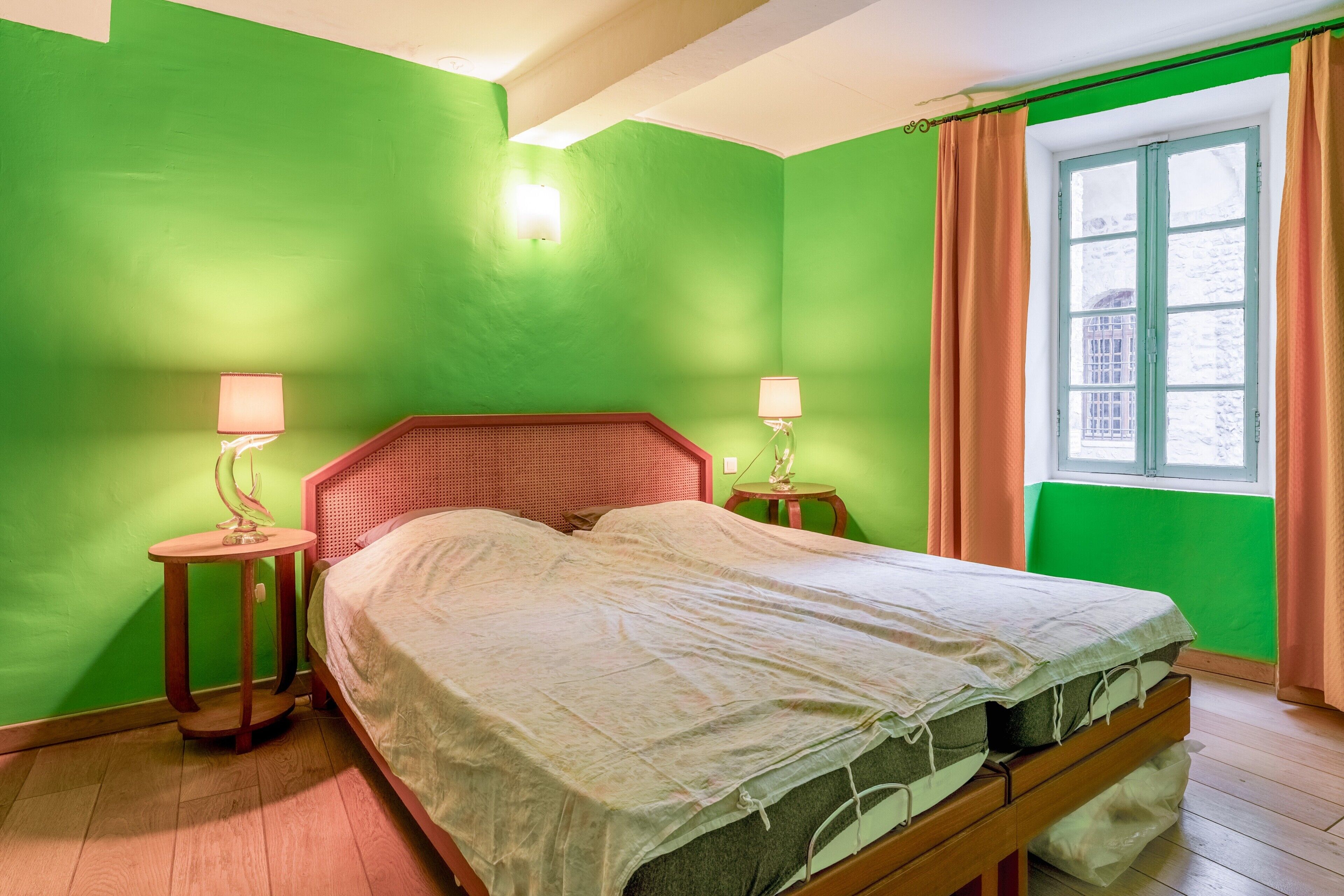 Una habitación con paredes verdes, una cama con cabecera de madera, dos mesitas de noche con lámparas y una ventana con cortinas.
