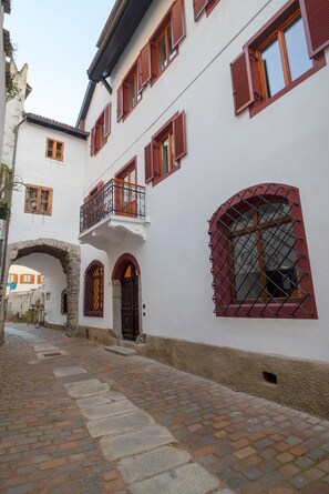 Une ruelle pavée étroite, bordée d’un bâtiment blanc aux volets rouges et surmontée d’une arche en pierre.