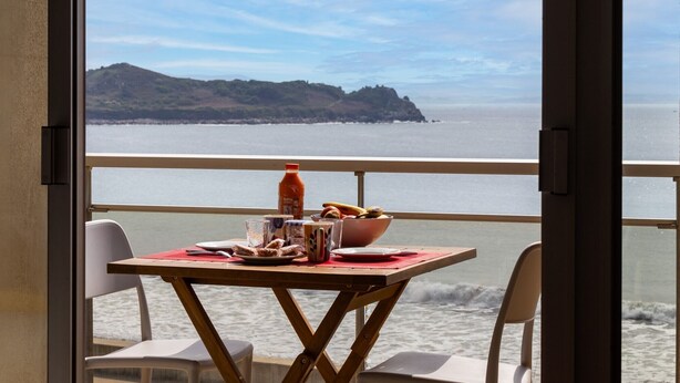 A balcony with a table set for two, overlooking a coastal view with a mountain in the distance. - Apartment 'Vue Mer By Madame Est Servie' with Sea View and Balcony (Trébeurden)