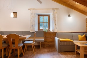 Una habitación acogedora con una mesa de madera, sillas, un sofá con cojines, una ventana con cortinas y una lámpara fijada a la pared.