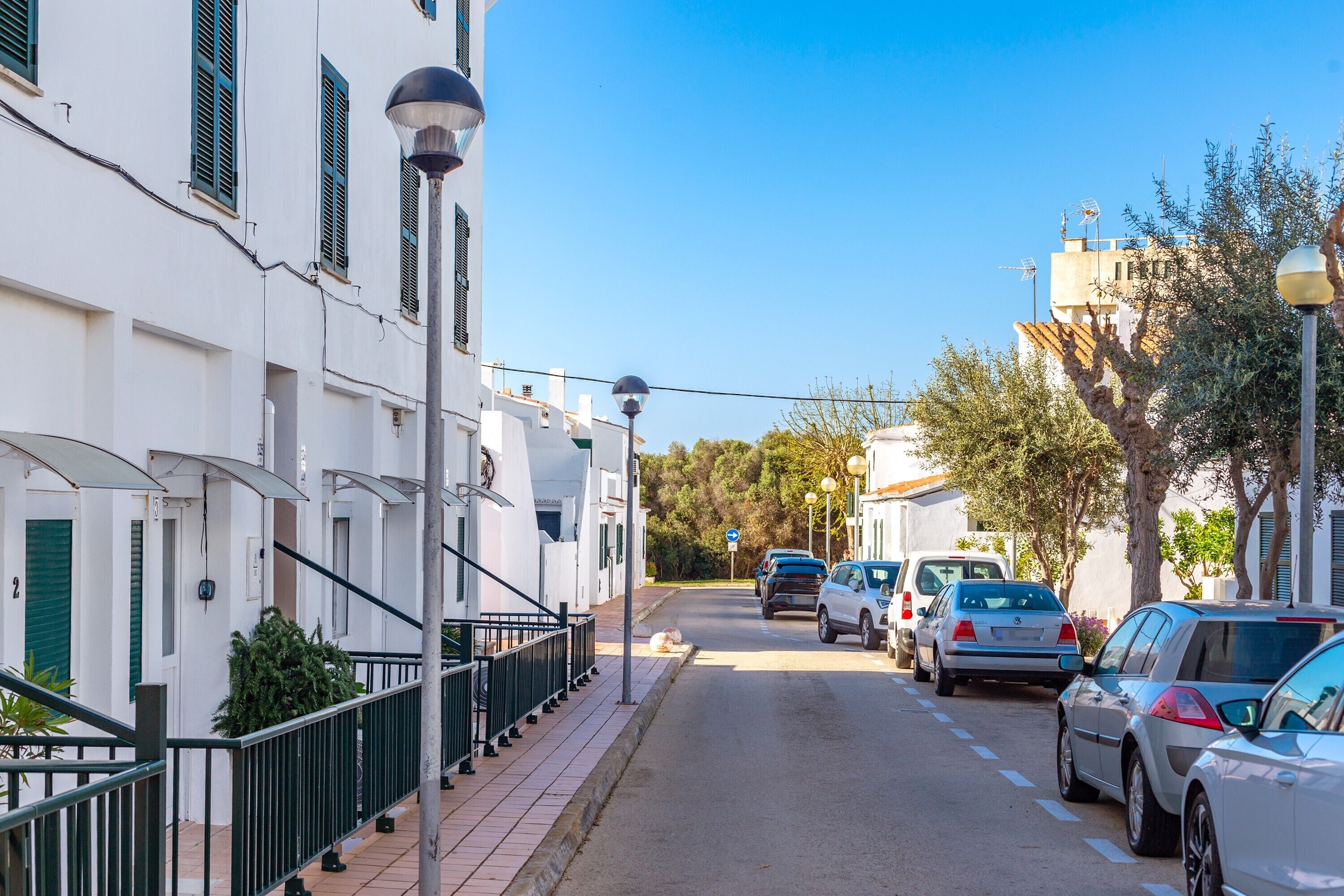 A narrow street lined with parked cars and white buildings with green shutters.