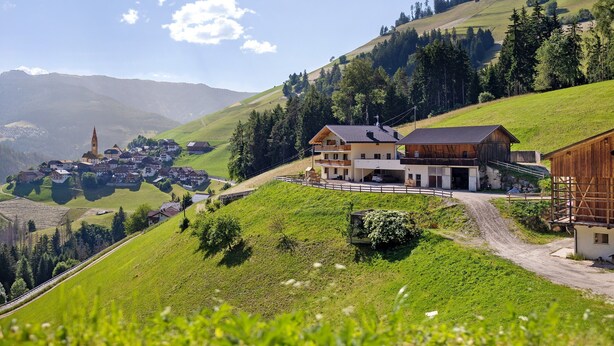 Un pittoresco borgo montano con una chiesa in evidenza, una grande casa di legno con un balcone e una strada sterrata che attraversa il villaggio.