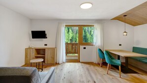 Una camera moderna con un tavolo da pranzo in legno, due sedie, un divano, una TV a parete e un balcone con vista sugli alberi.