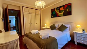 A hotel room with a bed, bedside lamps, a chair, a desk, a wardrobe, and a painting on the wall.