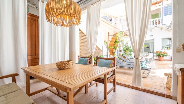 Un tavolo da pranzo in legno con sedie, un cesto intrecciato e un lampadario in una stanza luminosa con ampie finestre e tende.