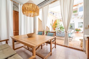 Una mesa de comedor de madera con sillas, una cesta tejida y una lámpara colgante en una habitación luminosa con ventanas grandes y cortinas.