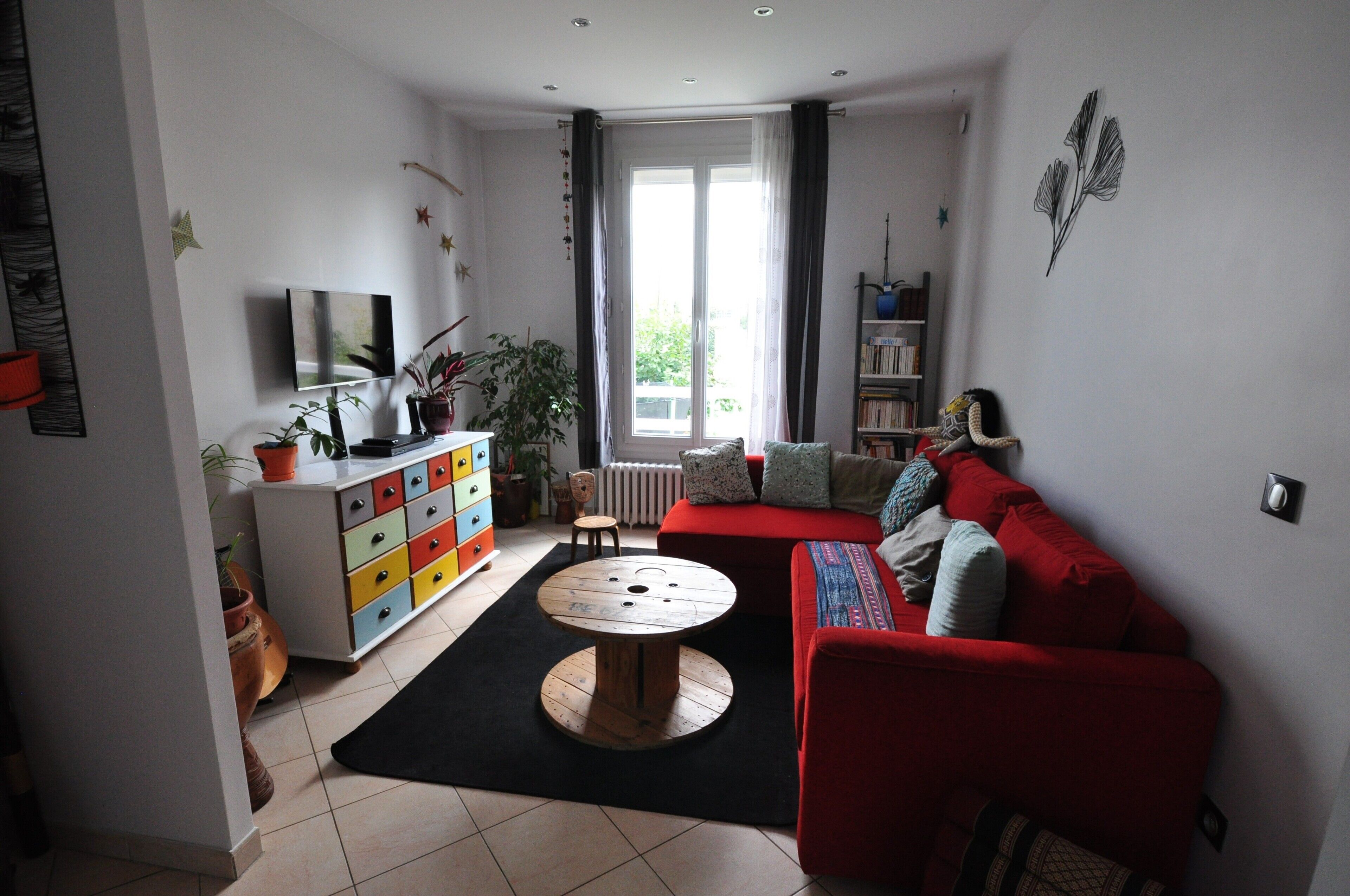 Un salon avec un canapé rouge, un téléviseur posé sur un meuble blanc, une table ronde en bois et une fenêtre avec des rideaux.