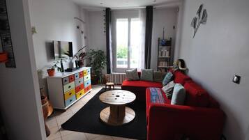 Un salon avec un canapé rouge, un téléviseur posé sur un meuble blanc, une table ronde en bois et une fenêtre avec des rideaux.