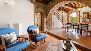 A rustic interior with exposed brick arches, wooden beams, a dining area with wooden chairs, and a blue upholstered chair with a cushion. - Holiday Home 'Toscana' with Shared Pool, Private Garden and Wi-Fi (Colle di Val d'Elsa)