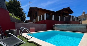 A swimming pool with a wooden deck, a metal ladder, and a red wall.