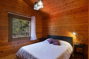 A wooden cabin room with a bed, a window with a curtain, a nightstand, and a lamp.