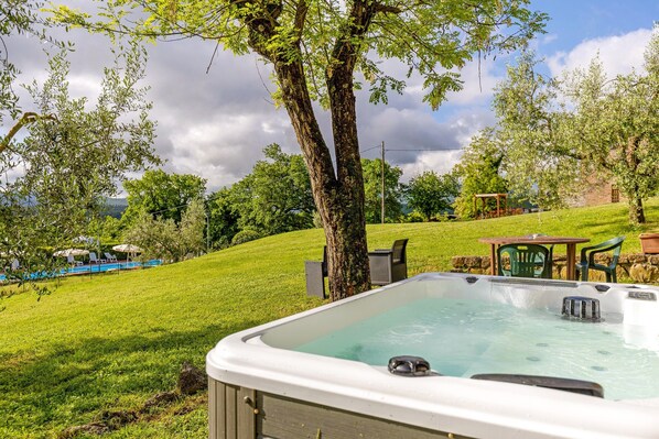 A hot tub in a backyard with a grassy lawn, trees, and a view of a swimming pool.