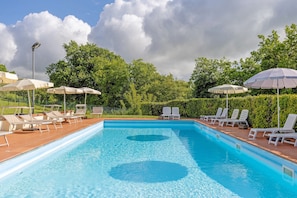A rectangular swimming pool with clear blue water, surrounded by white lounge chairs and umbrellas, set against a backdrop of lush green trees and a cloudy sky. - Cottage 'Nel Verde Con Jacuzzi' with Shared Pool and Shared Garden (Colle di Val d'Elsa)