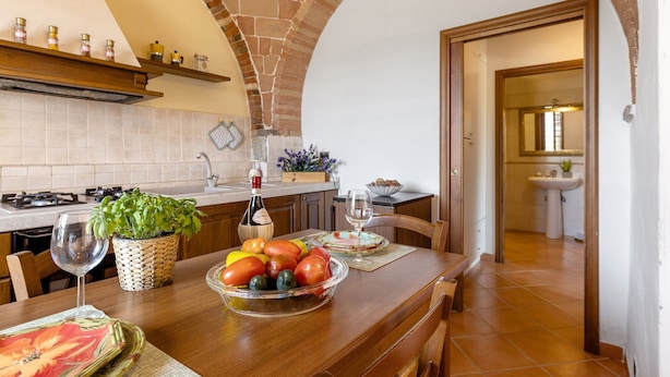 A kitchen with a brick archway, wooden cabinets, a dining table with a fruit bowl, a wine glass, and a bottle.