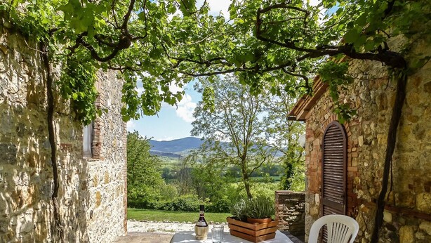 A stone building with a table and chairs, a potted plant, and a view of greenery and mountains.