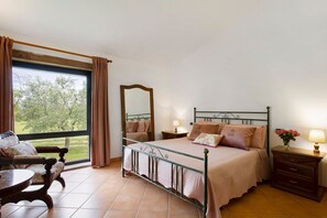 Een slaapkamer met een bed, een stoel, een tafel, een dressoir, een spiegel en een raam met gordijnen.