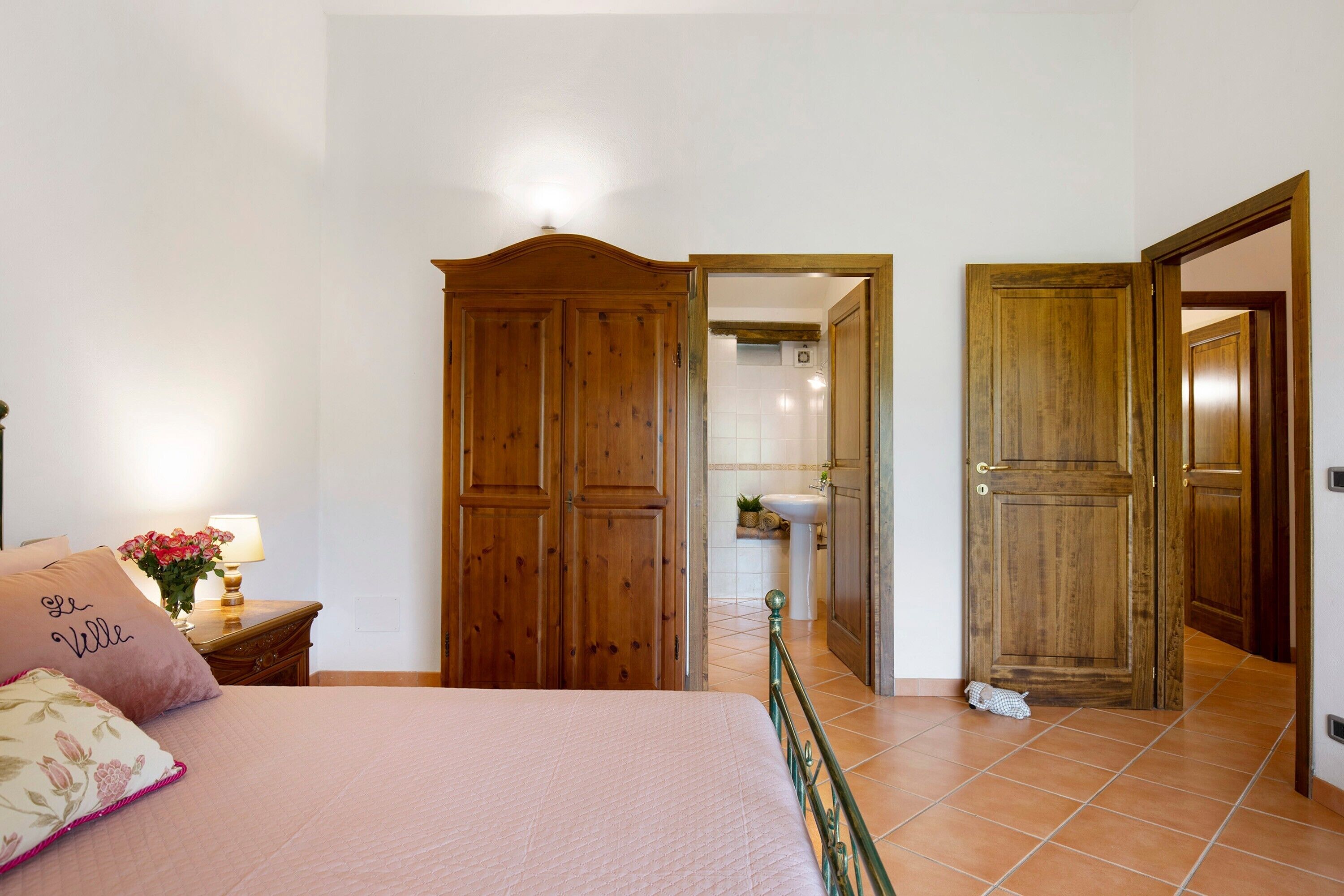 A bedroom with a bed, a nightstand with a lamp, a wooden wardrobe, and a bathroom visible through an open door.