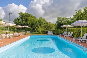 Een rechthoekig zwembad met helderblauw water, omringd door witte ligbedden en parasols, met op de achtergrond weelderige groene bomen en een bewolkte lucht.