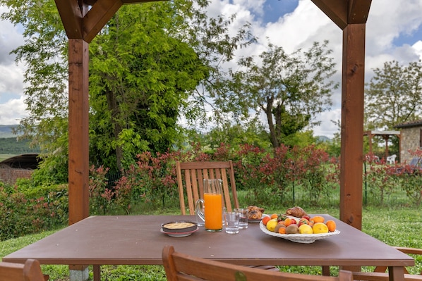 A wooden table with breakfast items, a pitcher of orange juice, and a chair in a garden setting.