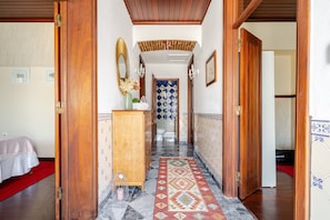 Un pasillo de hotel con una puerta de madera, un espejo, un jarrón con flores, un tapete estampado y un muro de azulejos.