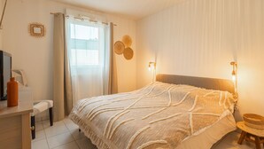 A bedroom with a bed, bedside lamps, a window with curtains, a wall-mounted TV, a small table with a chair, and decorative wall art. - Les Jardins d'Aunis by Interhome (Royan)