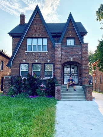 A two-story brick house with a steeply pitched roof, a front porch with a decorative arch, and a large front door with a wrought iron gate.