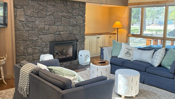 A cozy living room with a stone fireplace, a blue sofa, and a large window offering a view of the outdoors. - Meadow House 64 in Sunriver w/ Golf Course & Mt View, WiFi, AC, Hot Tub (Sunriver)
