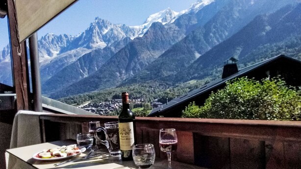 Ein Tisch mit einer Weinflasche, Gläsern und einem Teller mit Essen auf einem Balkon mit Bergblick.