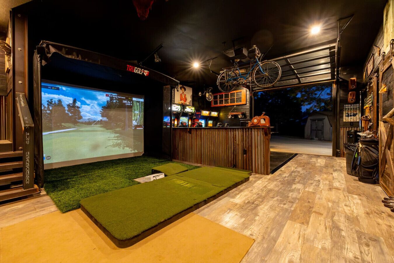 A room with a large screen displaying a golf game, a wooden bar area, and a bicycle mounted on the wall.