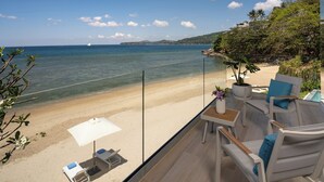 En balkon med udsigt over strand og hav, med et bord, stole og en potteplante.