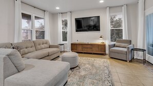 A modern living room with a sectional sofa, a grey armchair, a round ottoman, a wooden cabinet, a flat-screen TV, and a patterned rug. - Pet Friendly- Fort Morgan | Sleeps 12 | 3BR | 2.5BA (Gulf Shores)