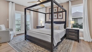 A four-poster bed with a dark frame, a bedside table with a lamp, and a view of the outside through a window with curtains. - Pet Friendly- Fort Morgan | Sleeps 12 | 3BR | 2.5BA (Gulf Shores)