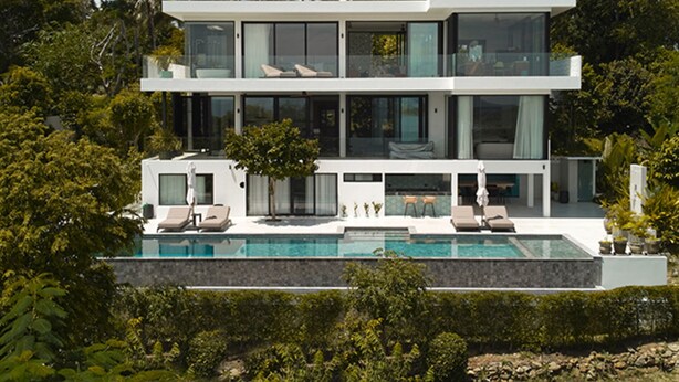 A modern two-story house with a swimming pool, surrounded by lush greenery and palm trees.