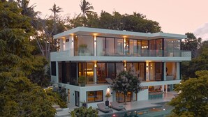 A modern two-story house with a glass facade, a swimming pool, and lush greenery surrounding it.