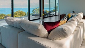 A modern living room with a large sofa, a red pillow, and a view of the sea through floor-to-ceiling windows.