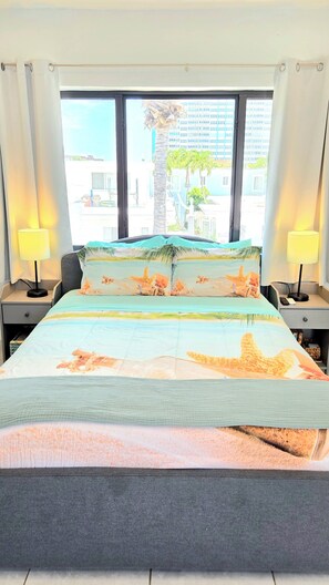 Una habitación de hotel con cama, dos mesitas de noche, ventana con cortinas y vistas a edificios y palmeras.