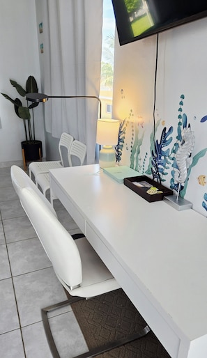 Una habitación moderna con un escritorio blanco, sillas, un televisor y un mural en la pared.