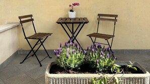 Un pequeño espacio exterior con dos sillas de madera y una mesa, una planta en maceta con flores moradas, y una lámpara fijada a la pared.