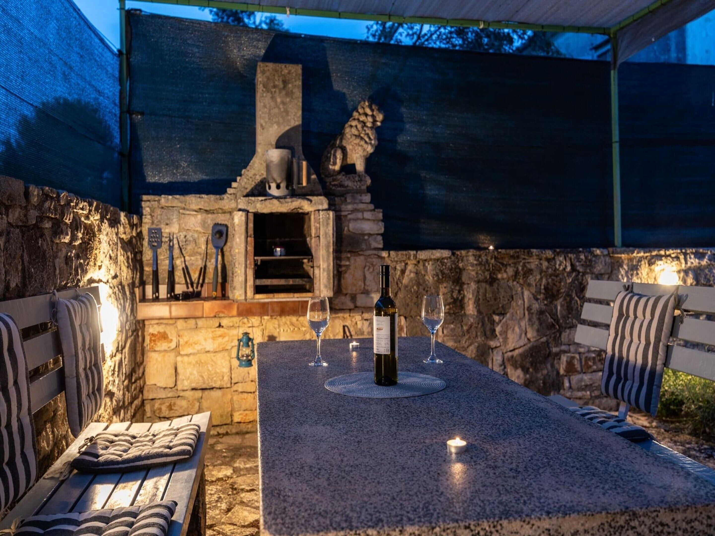 An outdoor patio with a stone fireplace, a table with wine glasses and a bottle, and striped cushions on benches.