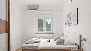 A bedroom with a wooden bed, a nightstand, a bedside table, a window with a view of greenery, and a wall-mounted air conditioner. - Apartment in Vodnjan With Pool & Jacuzzi (Vodnjan)
