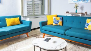 A modern living room with two blue sofas, yellow and blue patterned cushions, a white coffee table, and a painting on the wall.