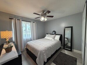 A bedroom with a bed, a ceiling fan, a bedside table with a lamp, a mirror, and a window with curtains.