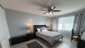 Un dormitorio con cama, dos mesitas de noche con lámparas, ventilador de techo, ventana con cortinas y una silla.