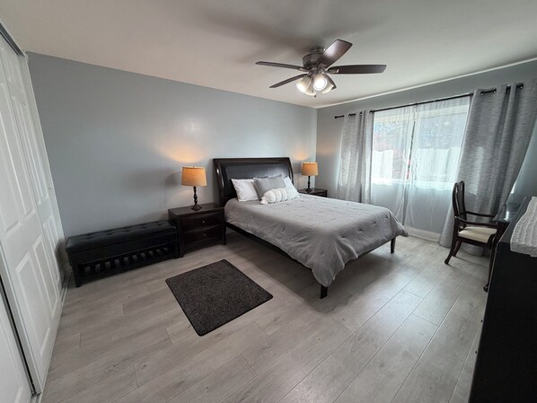 Un dormitorio con cama, dos mesitas de noche con lámparas, ventilador de techo, ventana con cortinas y una silla.