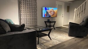 Een moderne woonkamer met een glazen salontafel, donkere banken, een kamerbreed gordijn en een flatscreen televisie op een standaard.