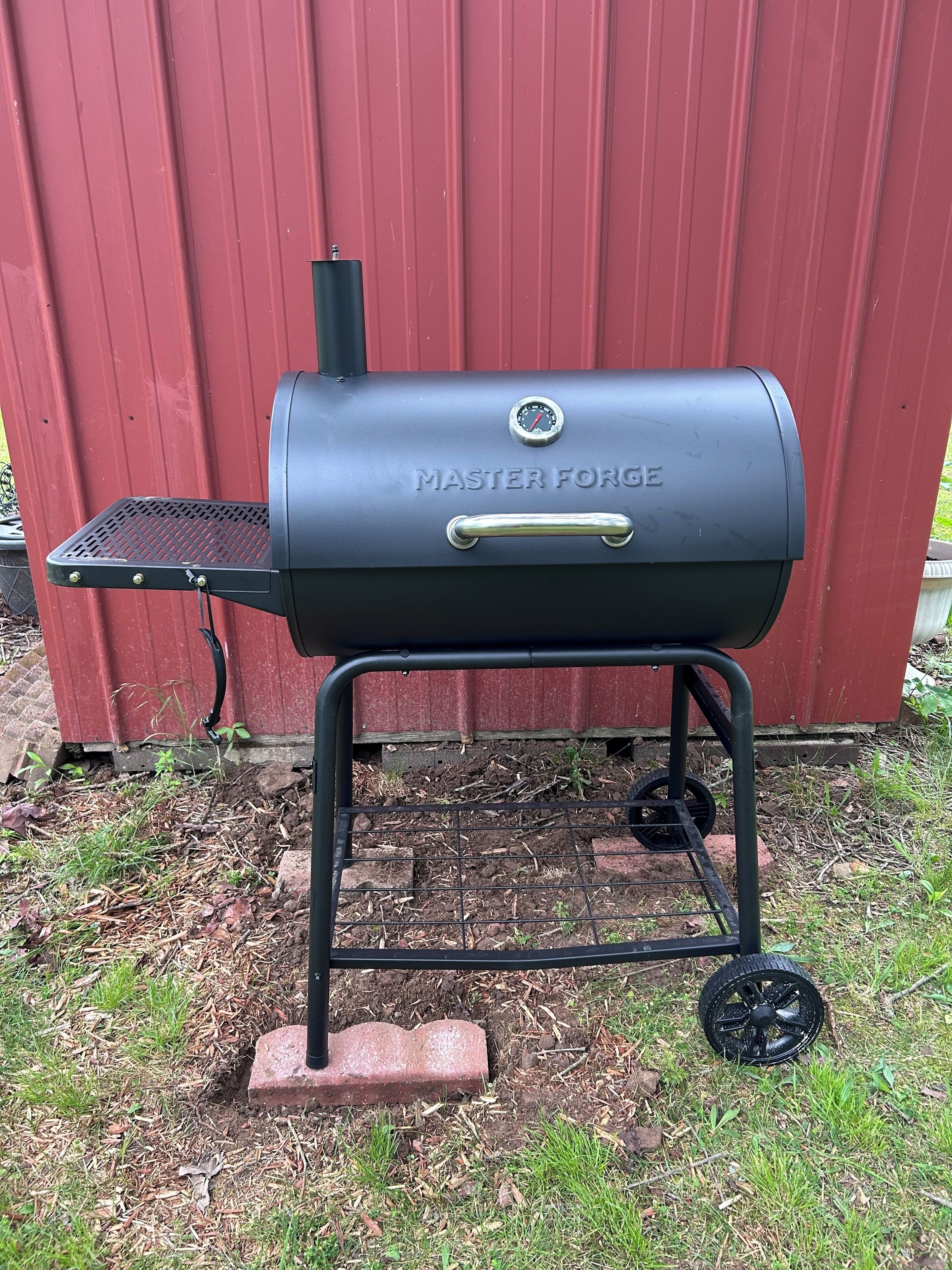 Een zwarte Master Forge barbecue met een bijzettafeltje, gemonteerd op een metalen onderstel met wielen, voor een rode achtergrond.